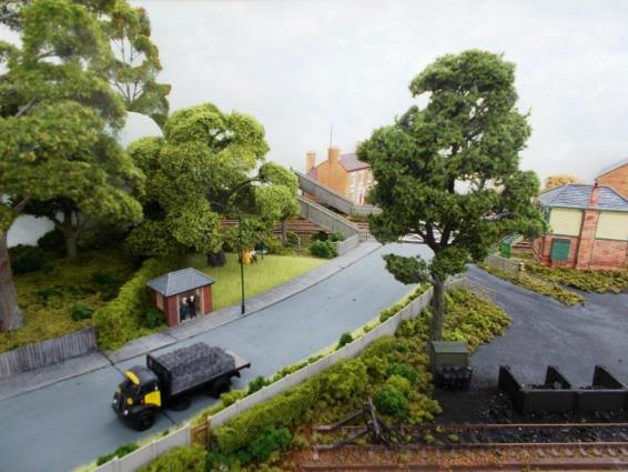 With its Mackenzie & Holland gates, Horsham Lane level crossing features a concrete footbridge which encroached into the front garden of Lavender House with its BBC H television ariel. To the left of the footbridge behind the bus stop a tree hides the scenic backboard with assists trains in disappearing from view. On the right part of the Signal Box is visible; this being now demoted to a Level Gate Crossing box. 
Below is virtually the same photograph shewing how a tree can significantly alter the view meaning the viewer now has to physically move to be able to see all.
www.EwhurstGreen.com 
