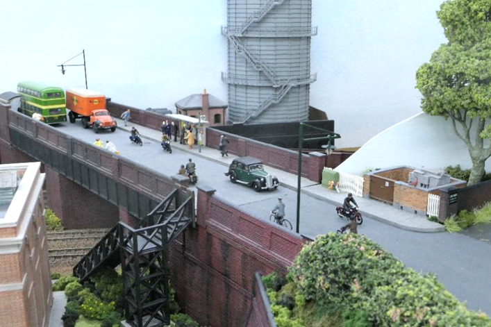 With the coming of the railway and associated development, the previously tranquil Somersbury Lane became a relatively busy street with its overbridge crossing both the Portsmouth main line and branch to Cranleigh at the country end of Ewhurst Green station.
In addition, a gas-holder (with gas supplied from Cranleigh gas works) became necessary (the Cranleigh branch is just visible in the foreground). Far from the lane’s rural setting, cycles, motor-cycles and motor vehicles cross the bridge as passengers wait for an omnibus.
www.EwhurstGreen.com 
