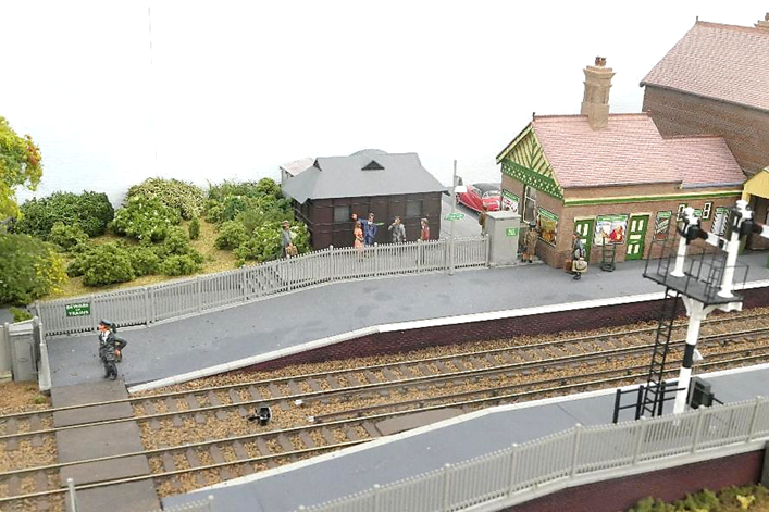 A model train station with a building and a small One Porter waits for a train to pass before using the barrow crossing while another struggles with a passengers luggage out to her red Austin Atlantic car.
A group of Teddyboys loiter outside the station café.
www.EwhurstGreen.com